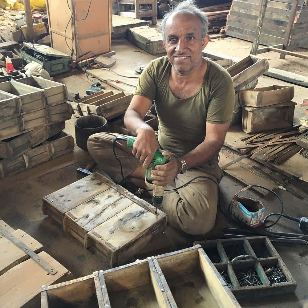 A man works with reclaimed wood, using an electric drill to assemble rustic wooden crates in a workshop filled with reclaimed materials and recycled furniture.